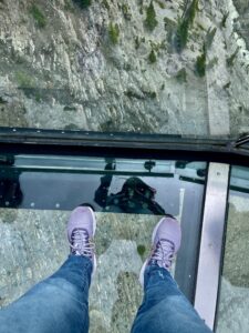 Person wearing purple shoes and jeans standing on a glass floor with a steep moss-covered cliff face visible below through the transparent surface
