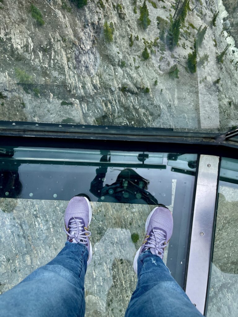 Person wearing purple shoes and jeans standing on a glass floor with a steep moss-covered cliff face visible below through the transparent surface
