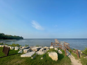 Wooden dock and rocky shoreline along a calm lake with forested peninsula in the distance under clear blue sky