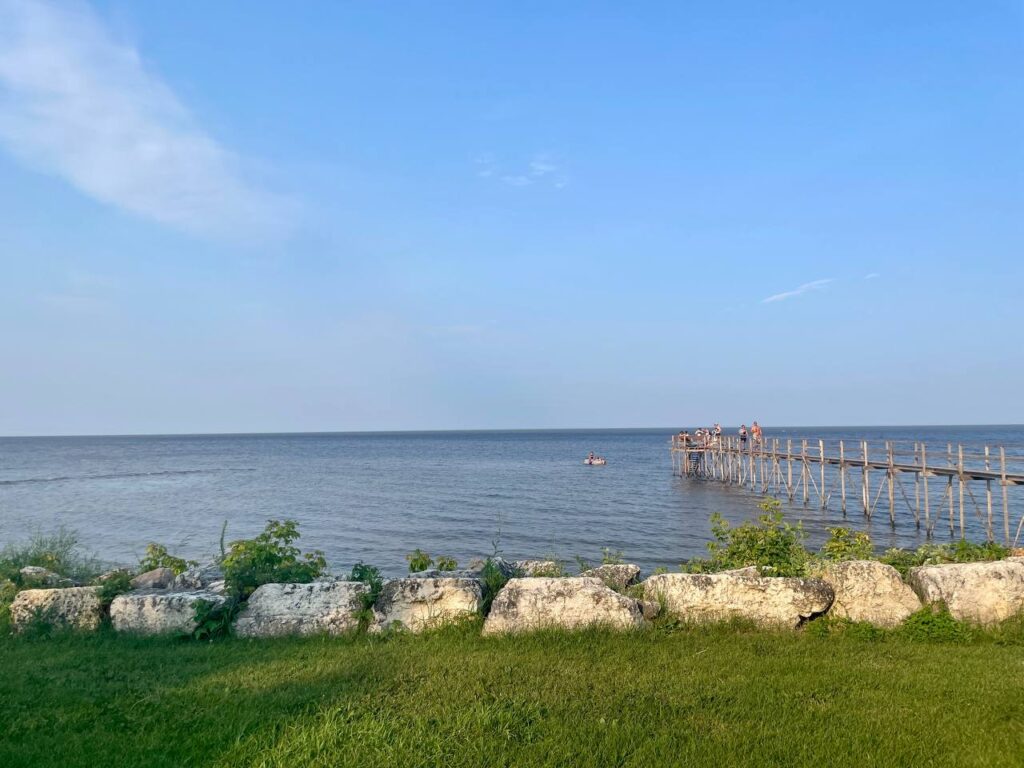 Waterfront view with wooden pier extending into calm water, stone seawall in foreground, green grass, and clear blue sky