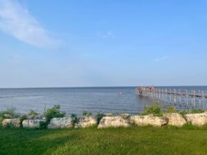 Waterfront view with wooden pier extending into calm water, stone seawall in foreground, green grass, and clear blue sky