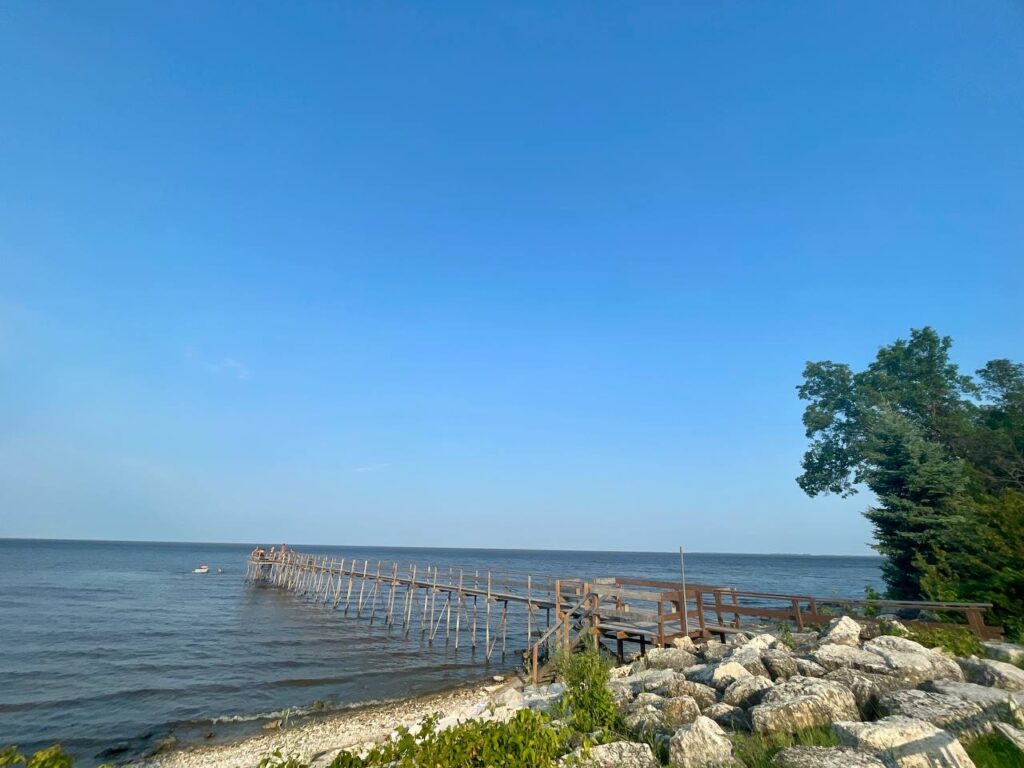 Weathered wooden pier extending into calm blue water, with rocky shoreline and green trees under clear sky