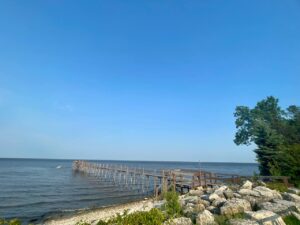 Weathered wooden pier extending into calm blue water, with rocky shoreline and green trees under clear sky