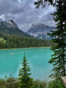 Turquoise alpine lake framed by coniferous forest, rocky mountains, and a red canoe on the water under cloudy sky