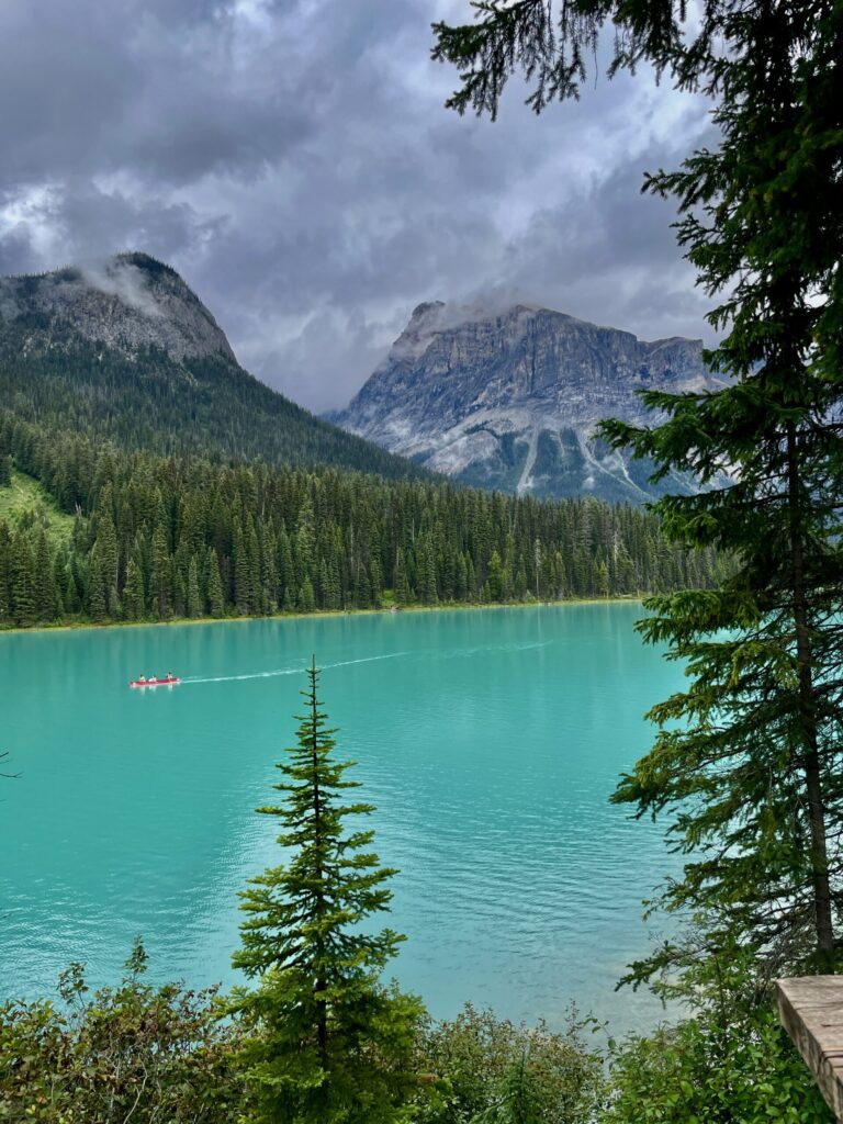 Turquoise alpine lake framed by coniferous forest, rocky mountains, and a red canoe on the water under cloudy sky