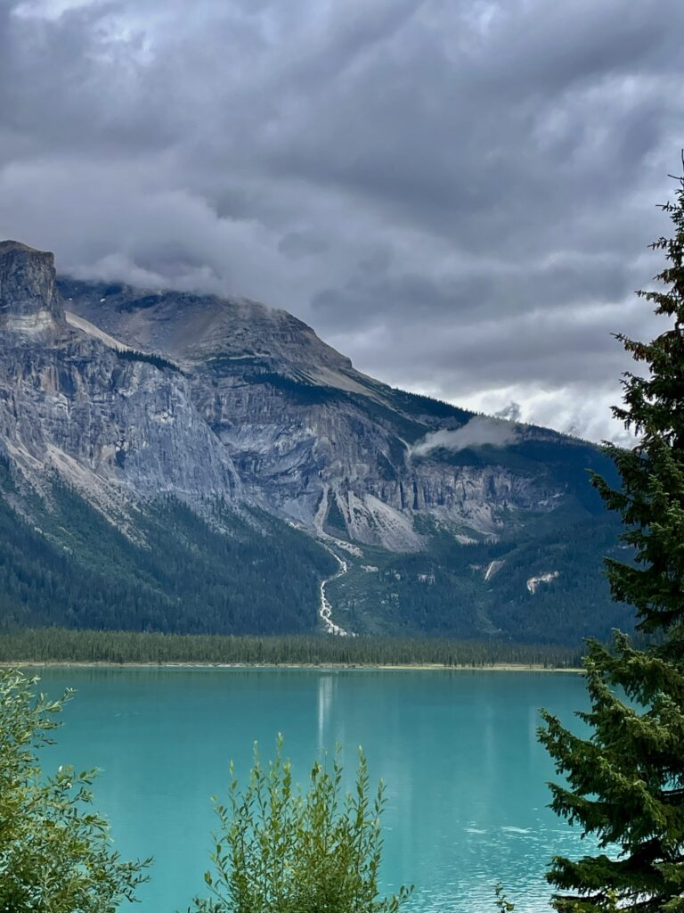 Turquoise alpine lake surrounded by coniferous forest, with dramatic rocky mountains and waterfalls under cloudy sky