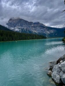 Turquoise alpine lake with rocky shoreline in foreground, dense coniferous forest along the far shore, and a dramatic layered mountain peak partially shrouded in cloud under an overcast sky