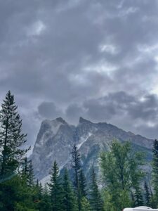 Dramatic rocky mountain peak with mist rising from its slopes, surrounded by coniferous and deciduous trees under a cloudy sky
