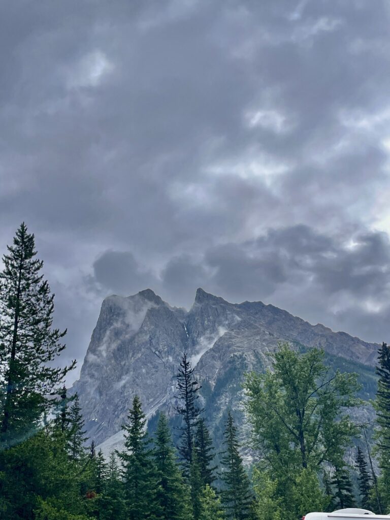 Dramatic rocky mountain peak with mist rising from its slopes, surrounded by coniferous and deciduous trees under a cloudy sky