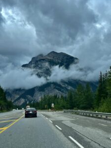 Mountain peak partially shrouded in mist rises above a highway lined with coniferous forest, with a red sedan driving ahead under overcast skies