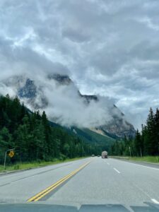 Straight highway through forested valley with dramatic rocky peaks shrouded in mist and low clouds, taken from inside a vehicle