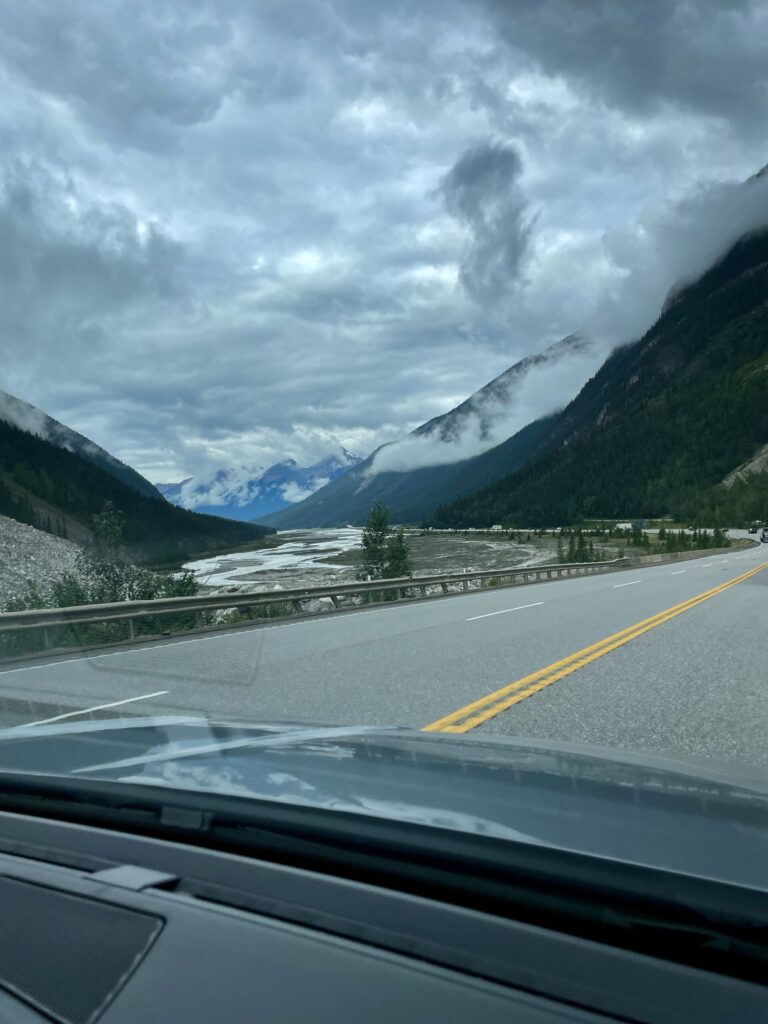 View from a vehicle crossing a bridge over a braided river valley, with forested mountains on both sides, snow-capped peaks in the distance, and dramatic cloudy sky overhead