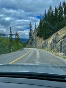 Mountain highway curving through rocky cliff face with coniferous forest, taken from inside a vehicle, with a large lake visible in the distance under a partly cloudy sky