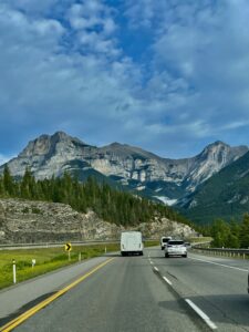 View from a highway with vehicles ahead, rocky mountains with layered cliff faces in the background, coniferous forest on the slopes, and a valley with mist below under a partly cloudy sky