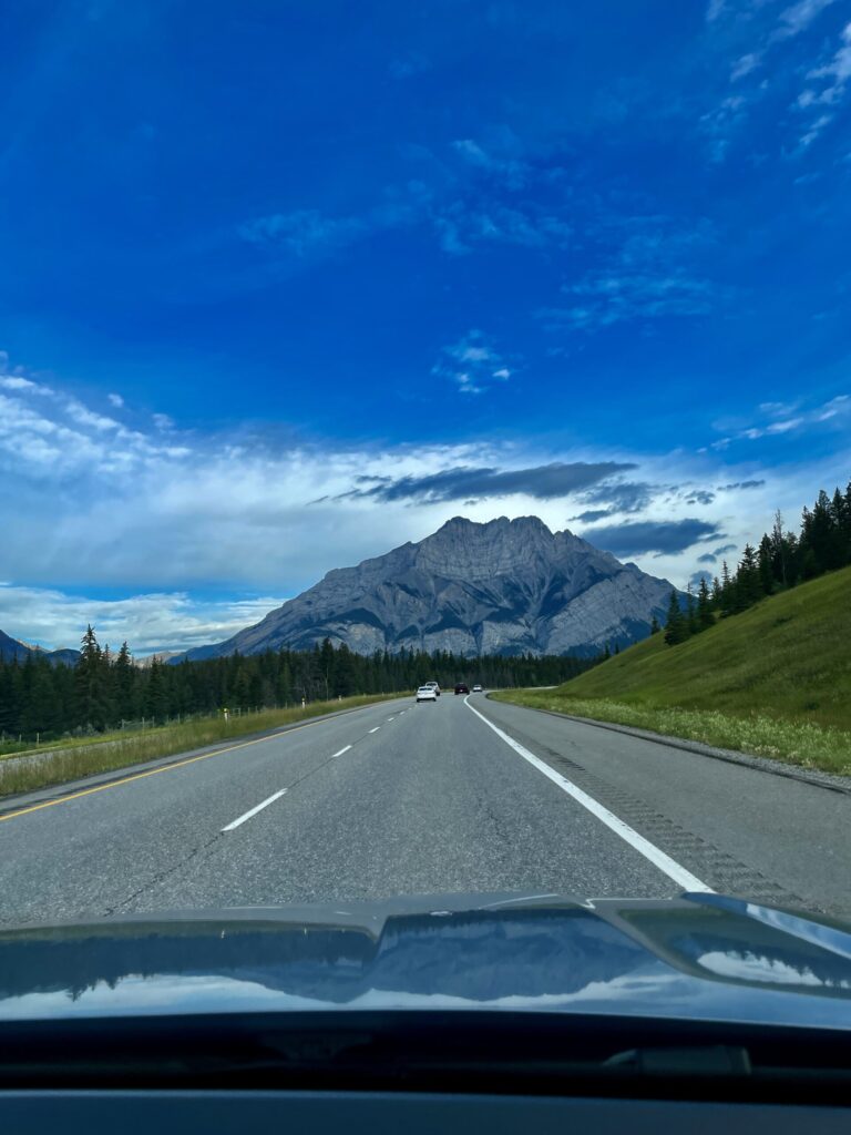 View from a vehicle driving on a highway toward a large rocky mountain peak, with coniferous forest on both sides, blue sky with clouds, and other vehicles ahead on the road