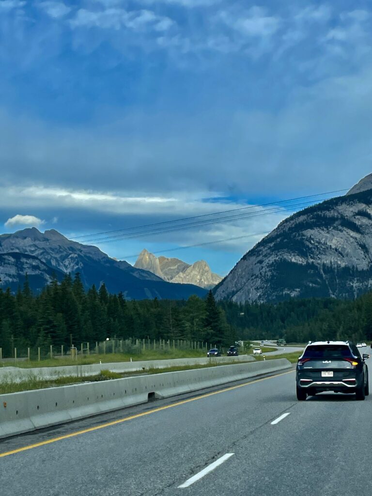 Highway driving through a mountain valley with coniferous forests, rocky peaks, and blue sky with clouds ahead