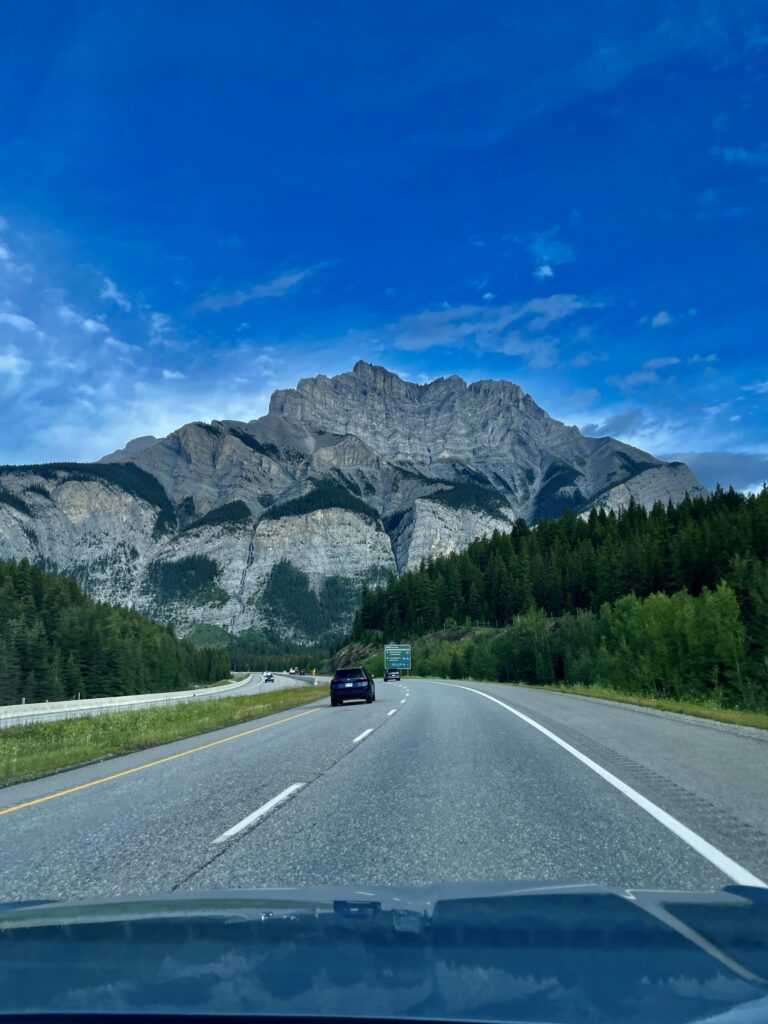 View from a vehicle driving on a highway toward a massive rocky mountain peak surrounded by coniferous forest under a clear blue sky