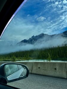View from inside a vehicle of a turquoise river, coniferous forest, and misty mountain peaks under a blue sky