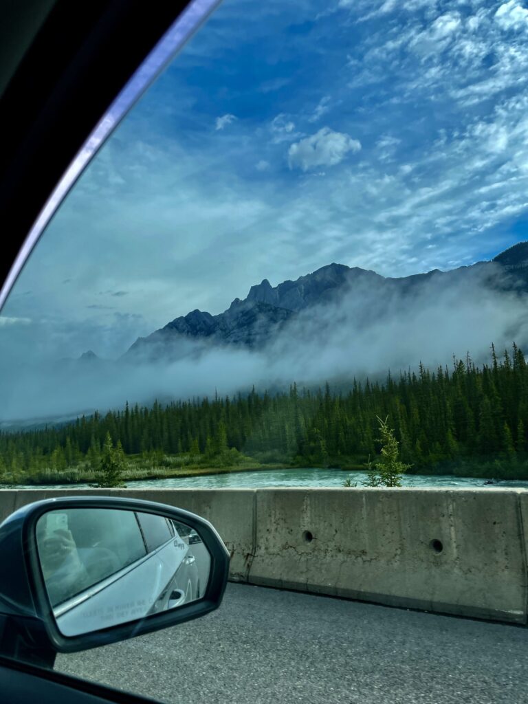 View from inside a vehicle of a turquoise river, coniferous forest, and misty mountain peaks under a blue sky