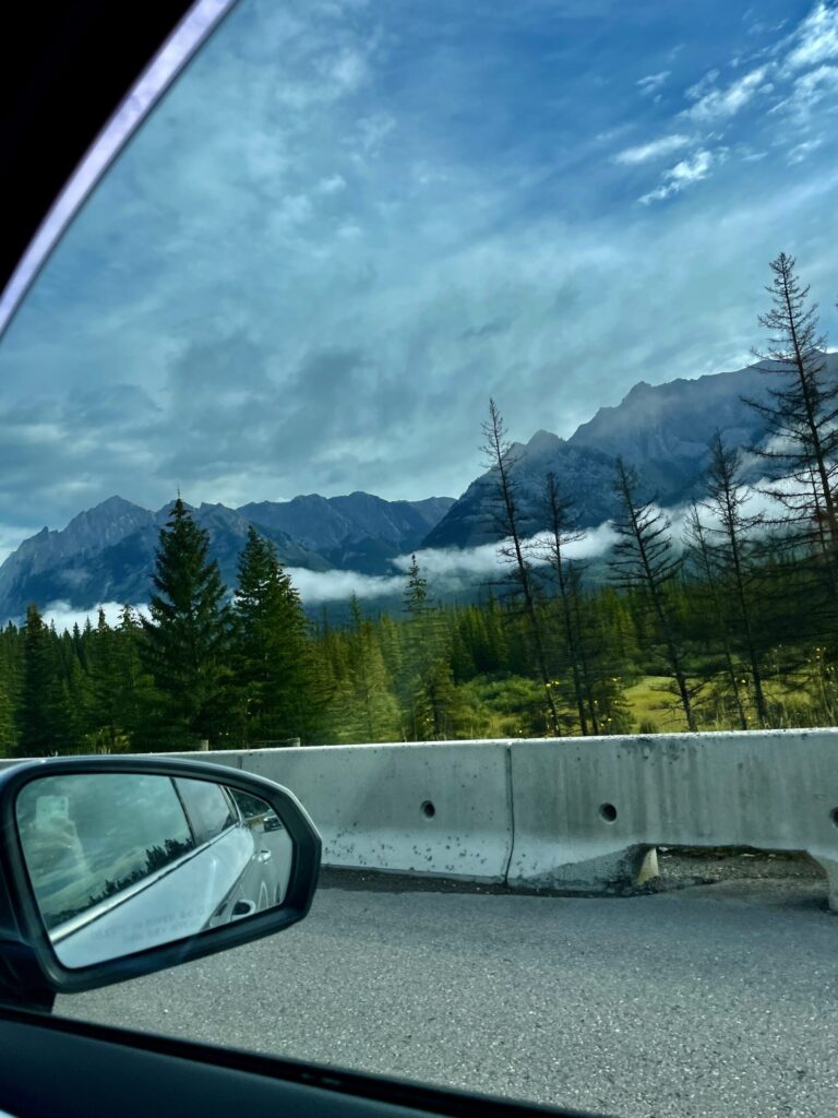 View from inside a vehicle of a mountain valley with coniferous forest, misty peaks, and blue sky, taken through the windshield and side mirror