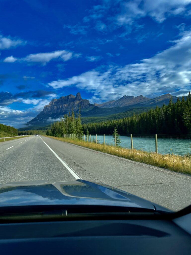 View from a vehicle driving along a highway beside a turquoise river, with a dramatic rocky mountain peak, coniferous forest, and blue sky with clouds in the distance