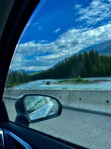 View from inside a vehicle showing a side mirror reflecting a turquoise river, coniferous forest, and mountain landscape under a blue sky with white clouds