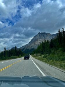 View from a vehicle driving on a mountain highway with a dark SUV ahead, coniferous forest on both sides, and a dramatic layered mountain peak under cloudy skies