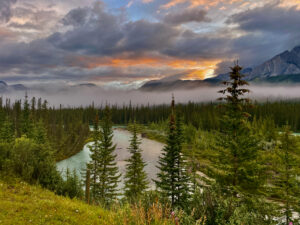 Mountain river winding through a coniferous forest valley at sunset, with dramatic clouds and peaks in the background
