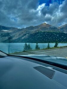 View from inside a vehicle during rainfall, looking through a rain-covered windscreen at a mountain lake surrounded by forested slopes and dramatic cloudy sky