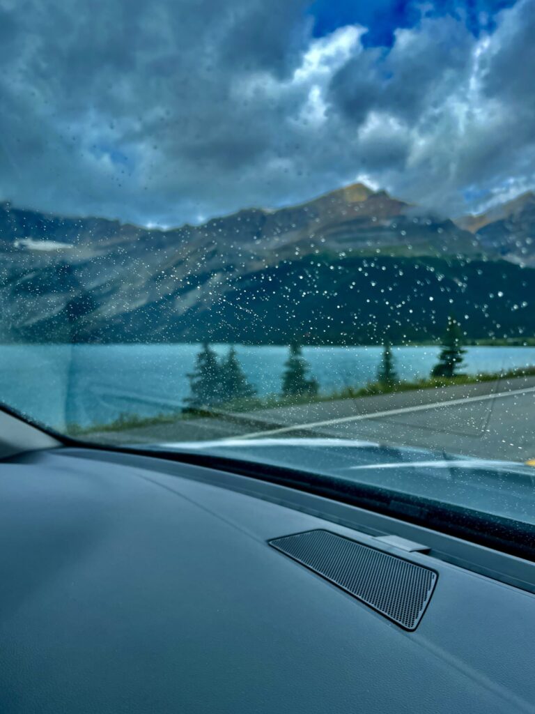 View from inside a vehicle during rainfall, looking through a rain-covered windscreen at a mountain lake surrounded by forested slopes and dramatic cloudy sky