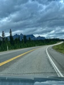 Highway curving through coniferous forest with jagged mountain peaks and dramatic storm clouds overhead