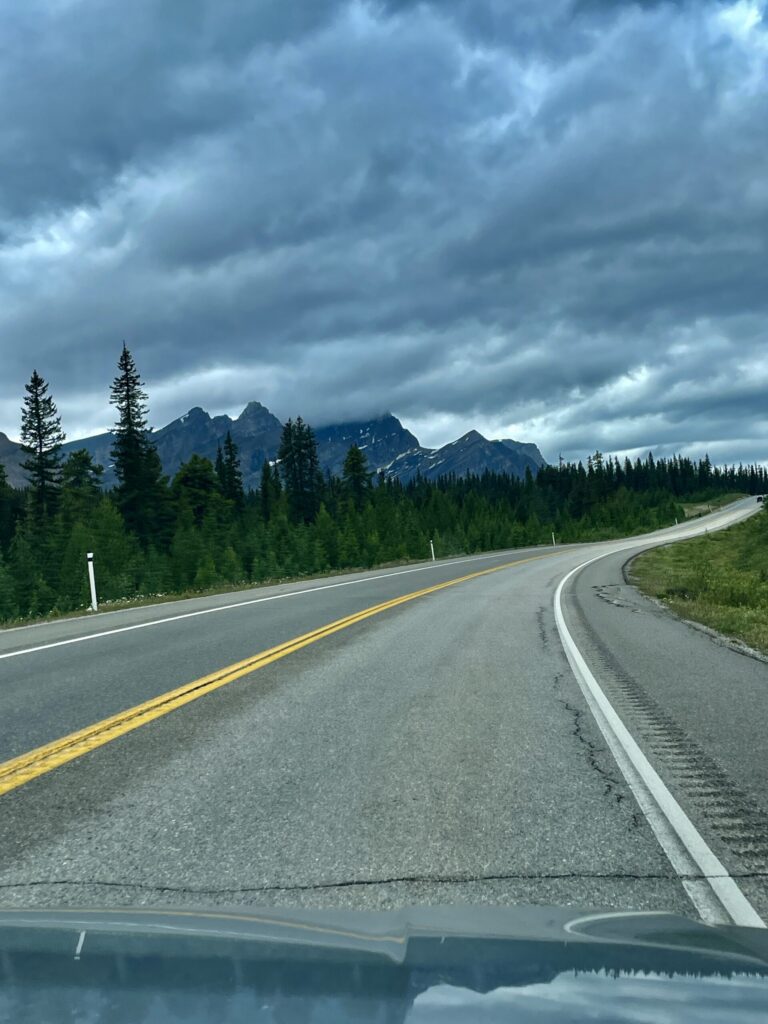 Highway curving through coniferous forest with jagged mountain peaks and dramatic storm clouds overhead