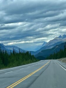 Straight highway with yellow centre line stretching toward snow-capped mountains, flanked by coniferous forest under dramatic cloudy sky