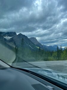 View from inside a vehicle of a mountain highway lined with coniferous forest, dramatic cloudy sky, and snow-capped peaks in the distance