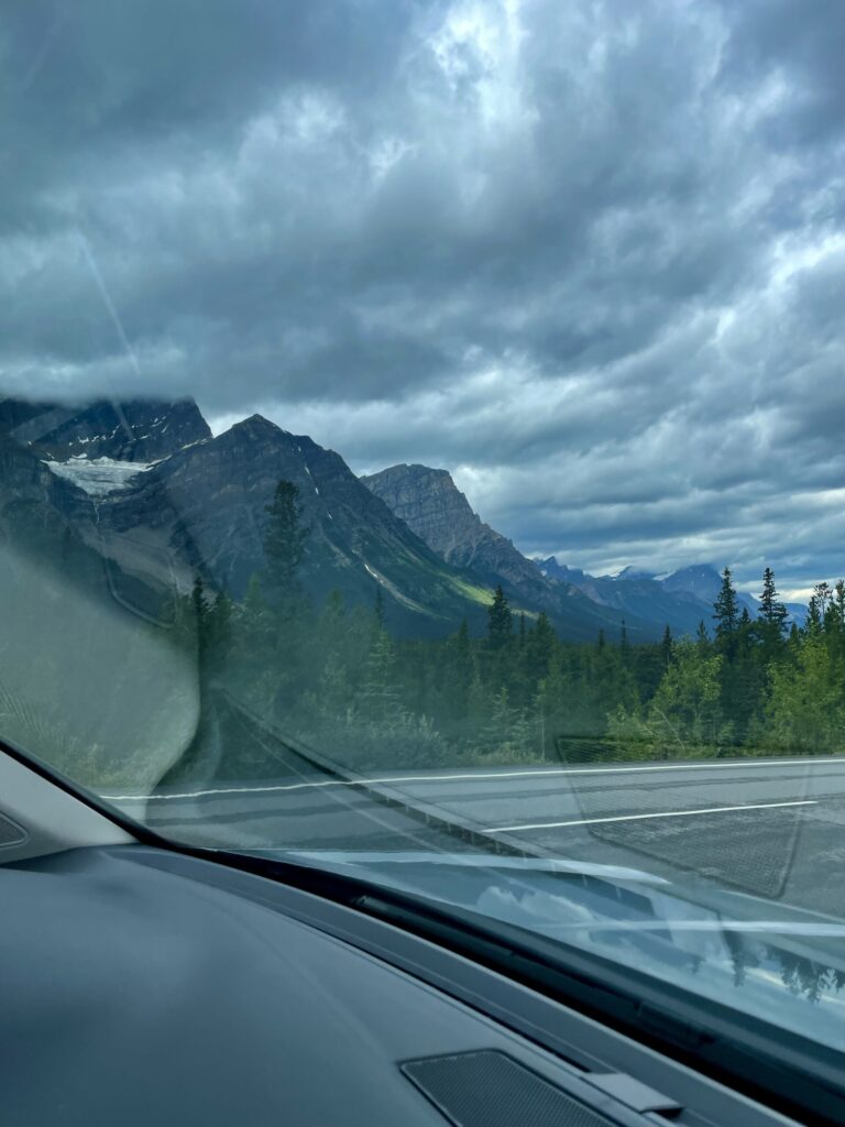 View from inside a vehicle of a mountain highway lined with coniferous forest, dramatic cloudy sky, and snow-capped peaks in the distance