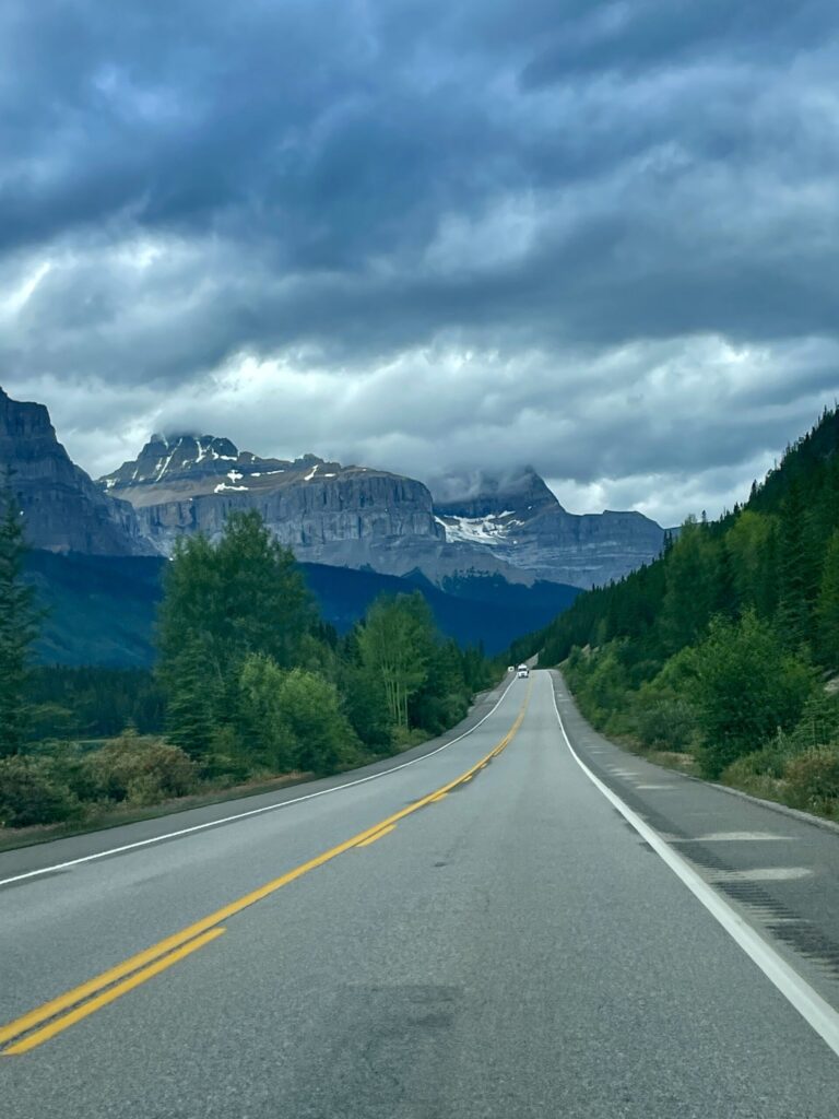 Straight highway stretching through a forested valley toward snow-capped mountains under dramatic cloudy skies