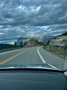 View from a vehicle driving on a mountain highway with yellow centre lines, curving past rocky outcrops, coniferous forests, and distant peaks beneath dramatic storm clouds