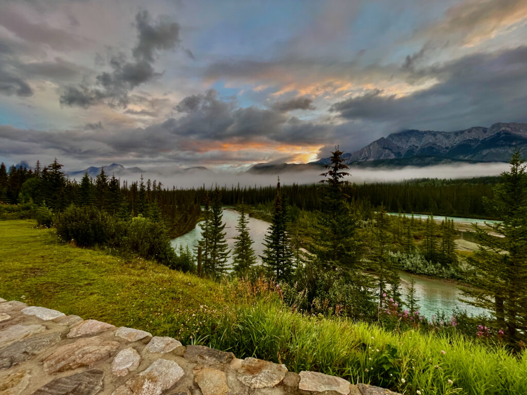Mountain valley with turquoise river winding through coniferous forest at sunset, dramatic clouds illuminated above snow-capped peaks