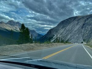 Highway stretching through a mountain valley with steep rocky peaks on either side, coniferous trees, and dramatic cloudy sky