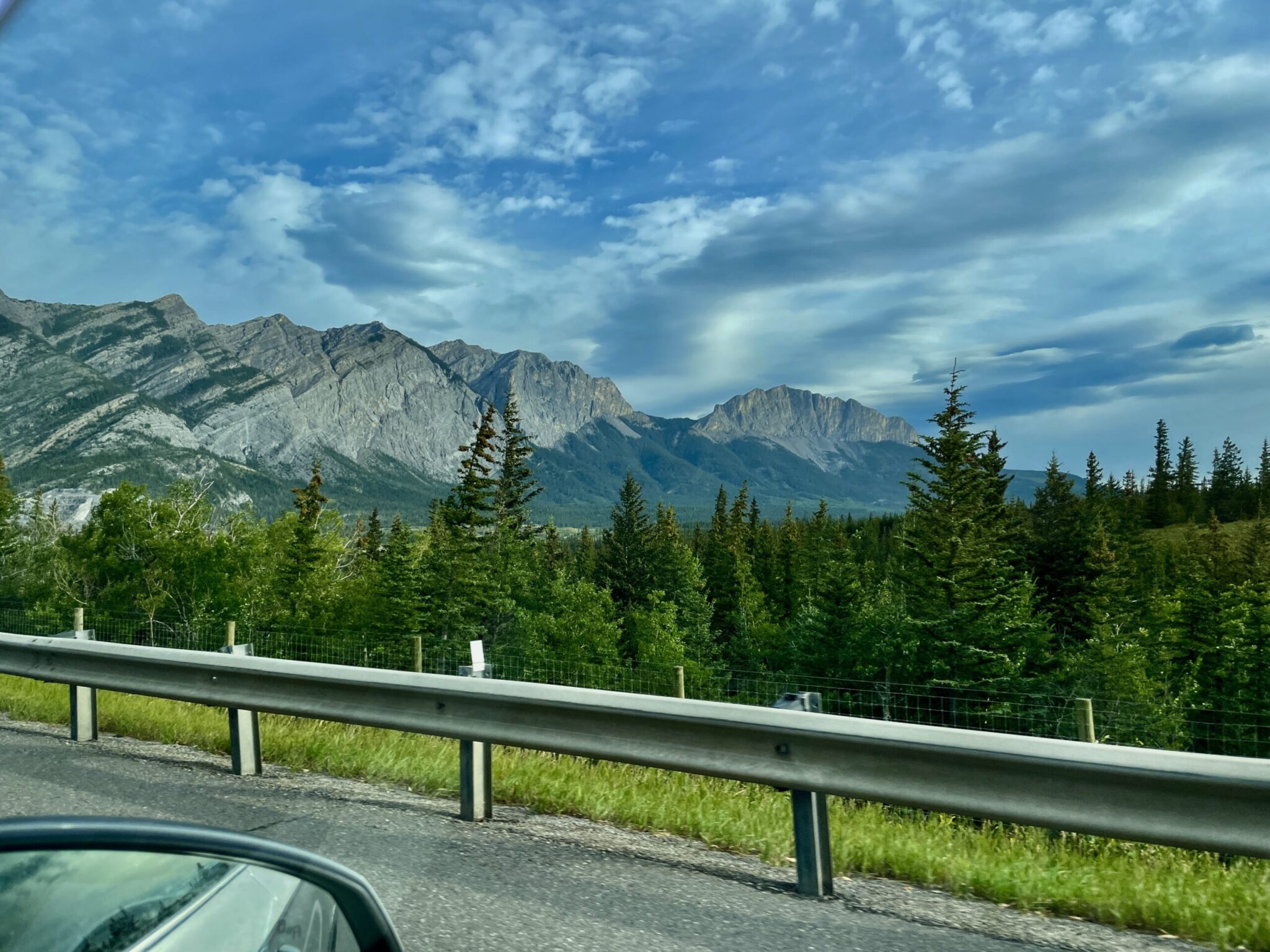 Mountain landscape with coniferous forest and layered peaks viewed from a roadside overlook with guardrail, under blue sky with white clouds