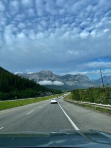 View from a vehicle driving on a highway through a mountain valley with snow-capped peaks, coniferous forest, and other vehicles ahead on the road