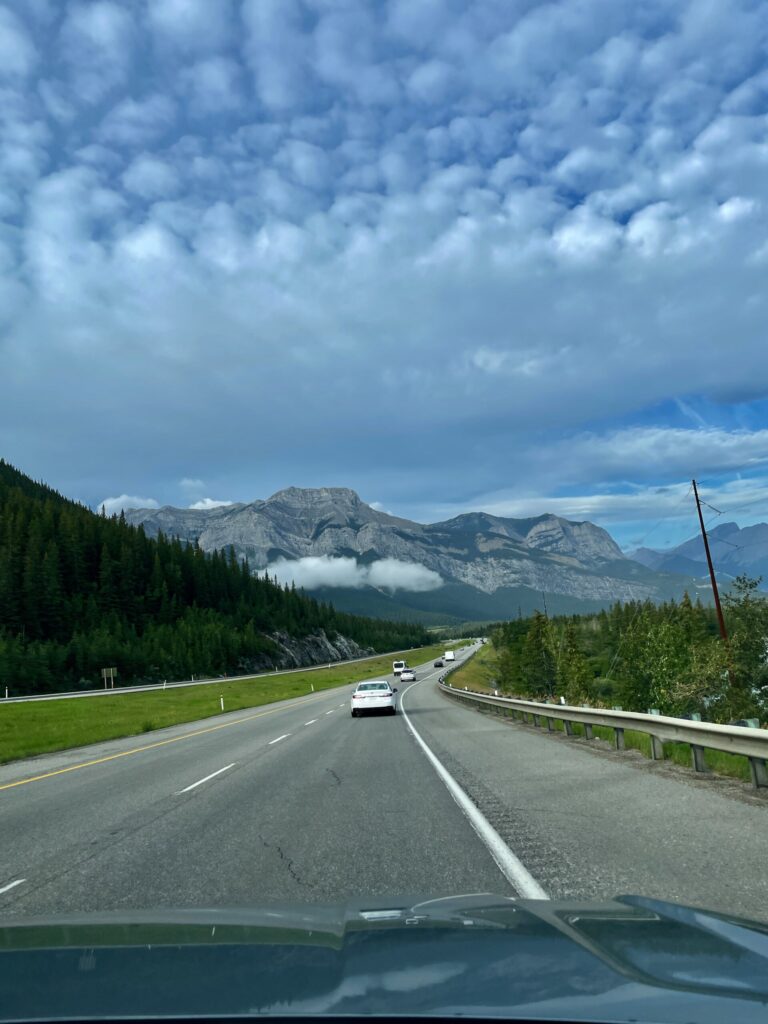 View from a vehicle driving on a highway through a mountain valley with snow-capped peaks, coniferous forest, and other vehicles ahead on the road