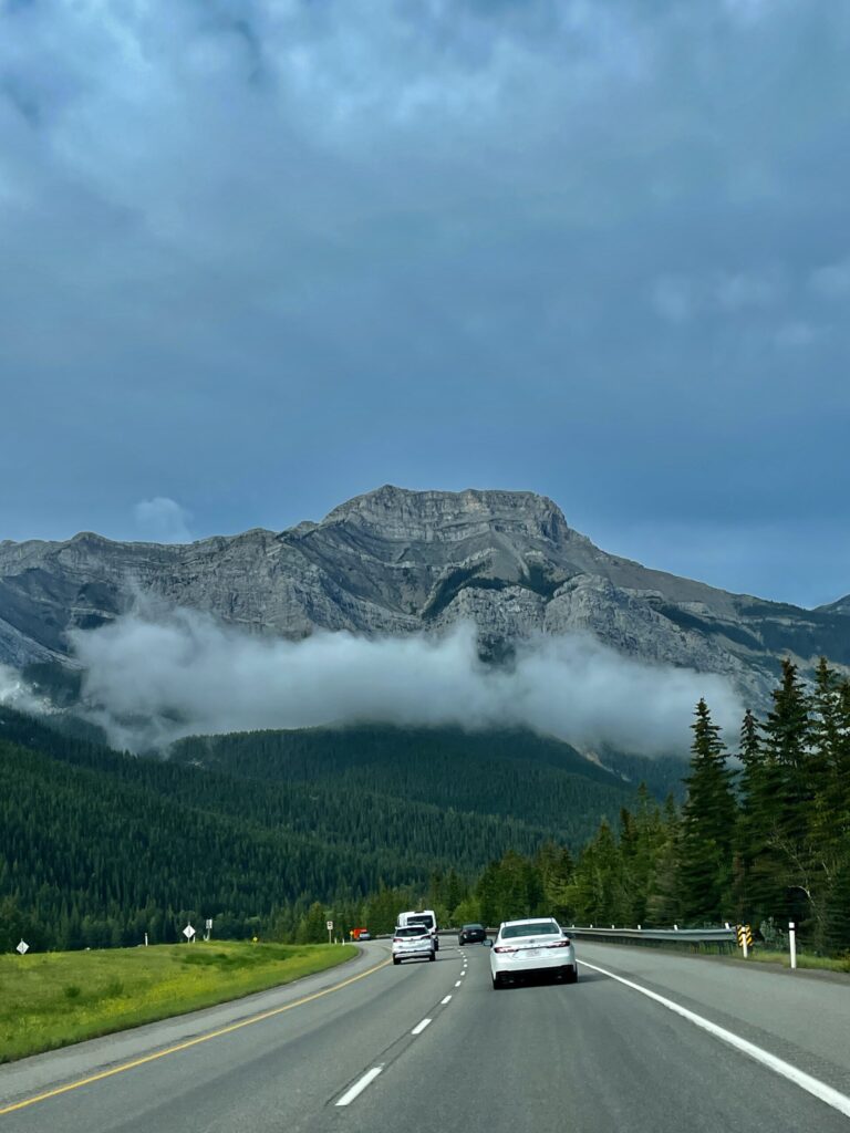 Highway stretching toward a dramatic mountain peak partially shrouded in mist, with dense coniferous forest and vehicles on the road