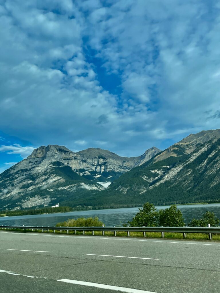 Mountain lake view from a highway with rocky peaks, coniferous forest, and blue sky with clouds