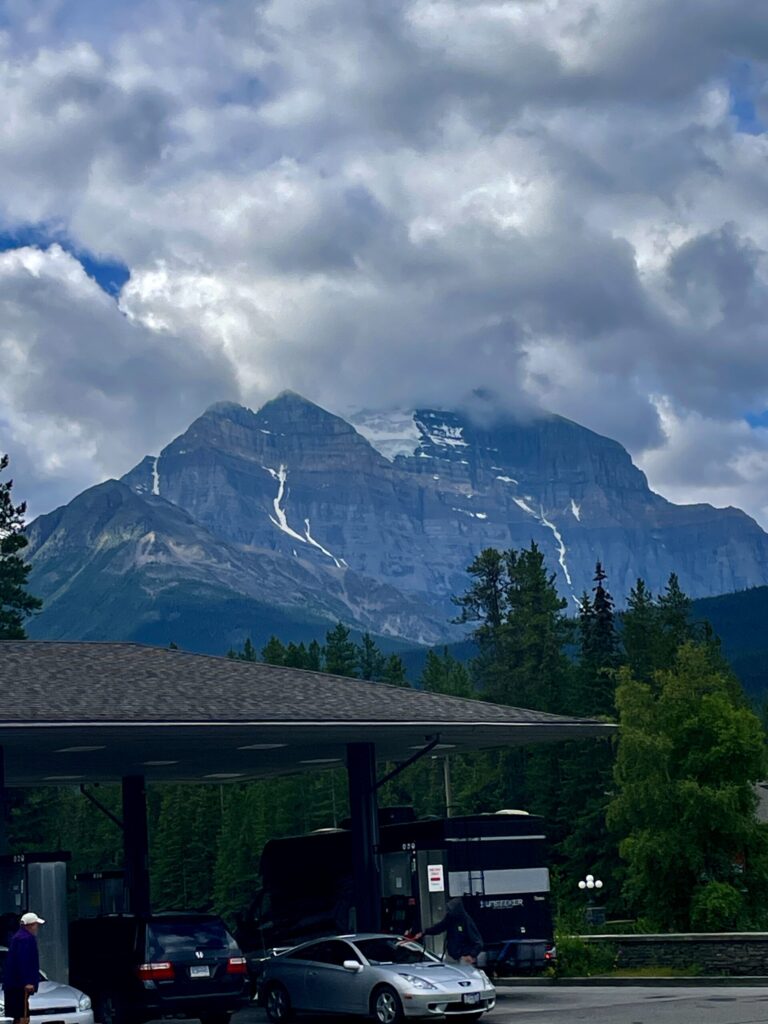 Snow-capped mountain with waterfalls visible on its face, framed by dramatic cloudy sky, viewed from a service station with parked vehicles in the foreground