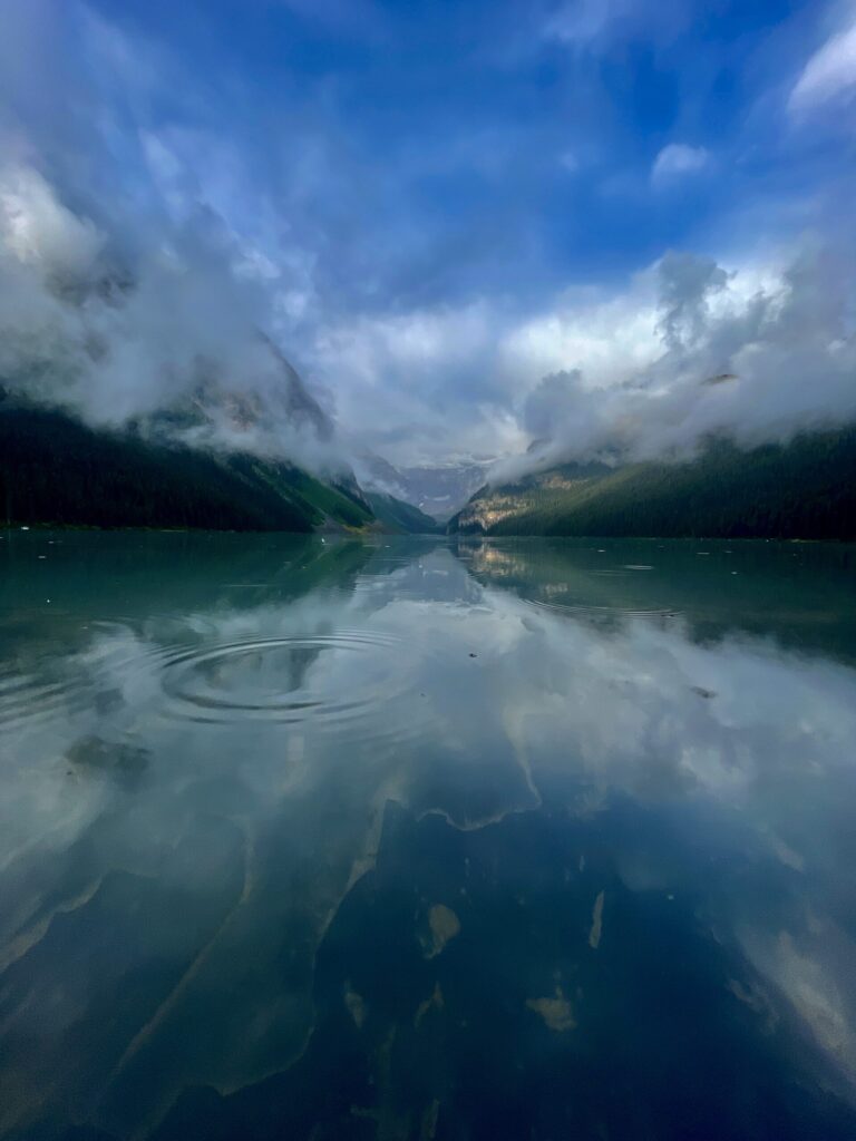 Turquoise alpine lake with misty mountains and forested shores, ripples spreading across calm water under a blue sky with white clouds