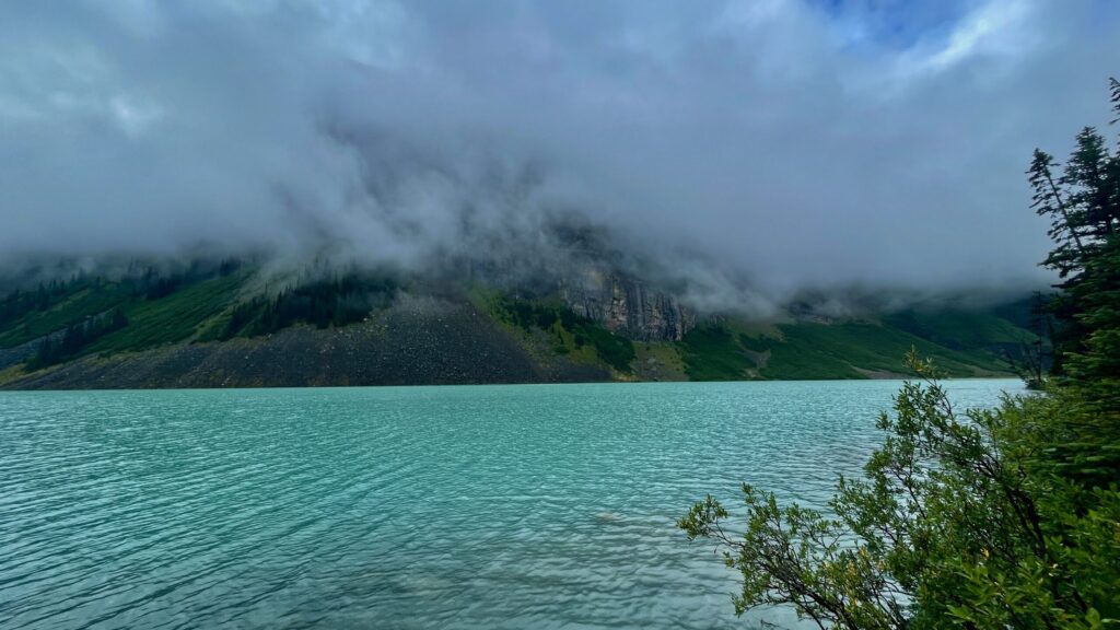 Turquoise alpine lake with forested mountains shrouded in mist and low clouds, rocky slopes visible through the fog