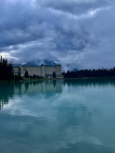 Large historic hotel on the shore of a turquoise lake, surrounded by coniferous forest, with snow-capped mountains and dramatic clouds in the background