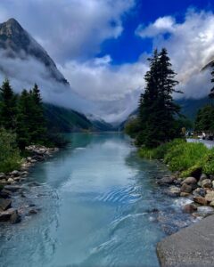 Turquoise alpine lake surrounded by coniferous forests, rocky shores, and mist-shrouded mountains under a partly cloudy sky