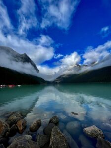 Turquoise alpine lake surrounded by forested mountains with mist-covered peaks under a blue sky, red canoes moored on the left shore